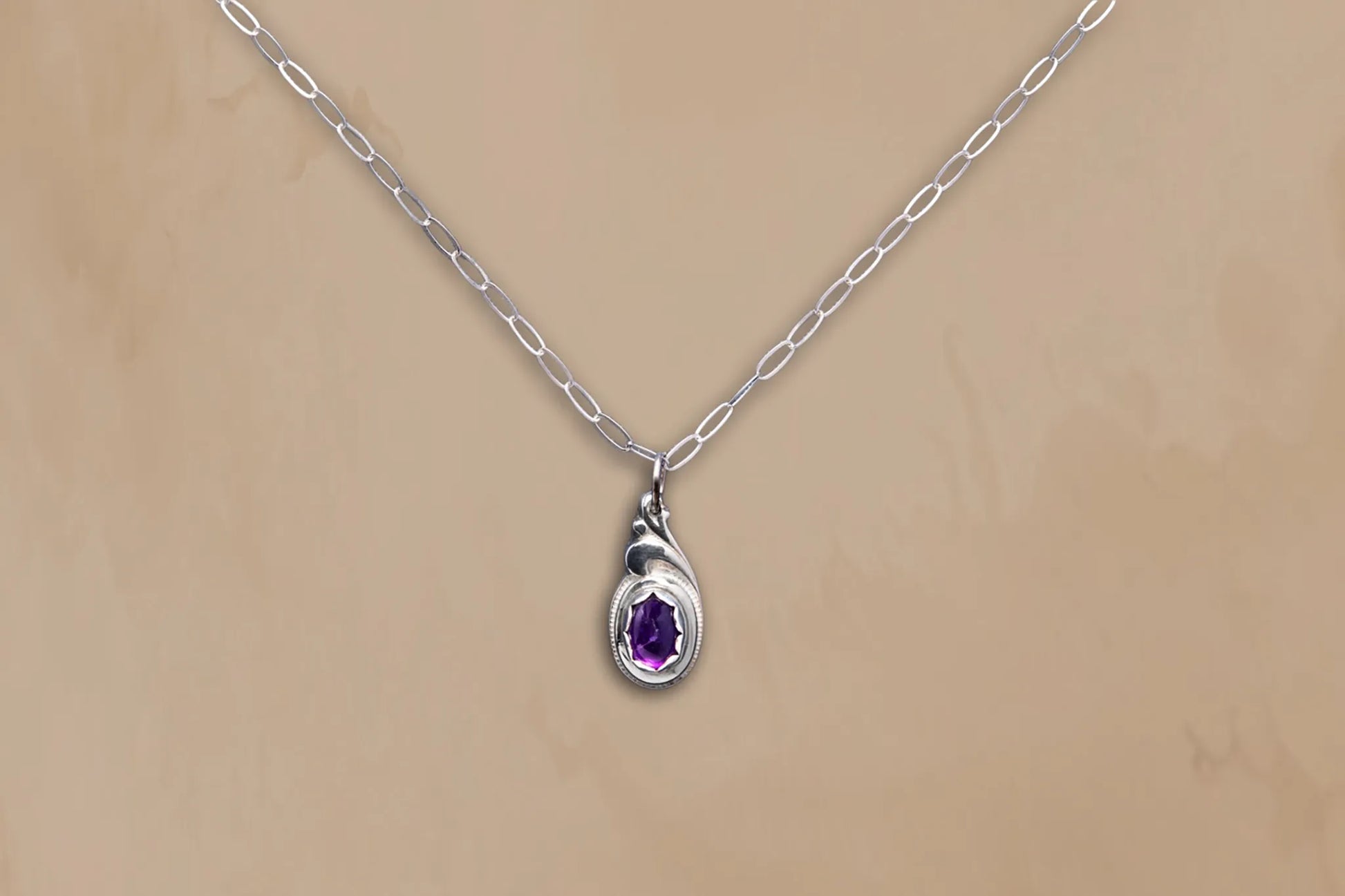 Silver necklace with a teardrop-shaped pendant featuring a purple gemstone on a beige background