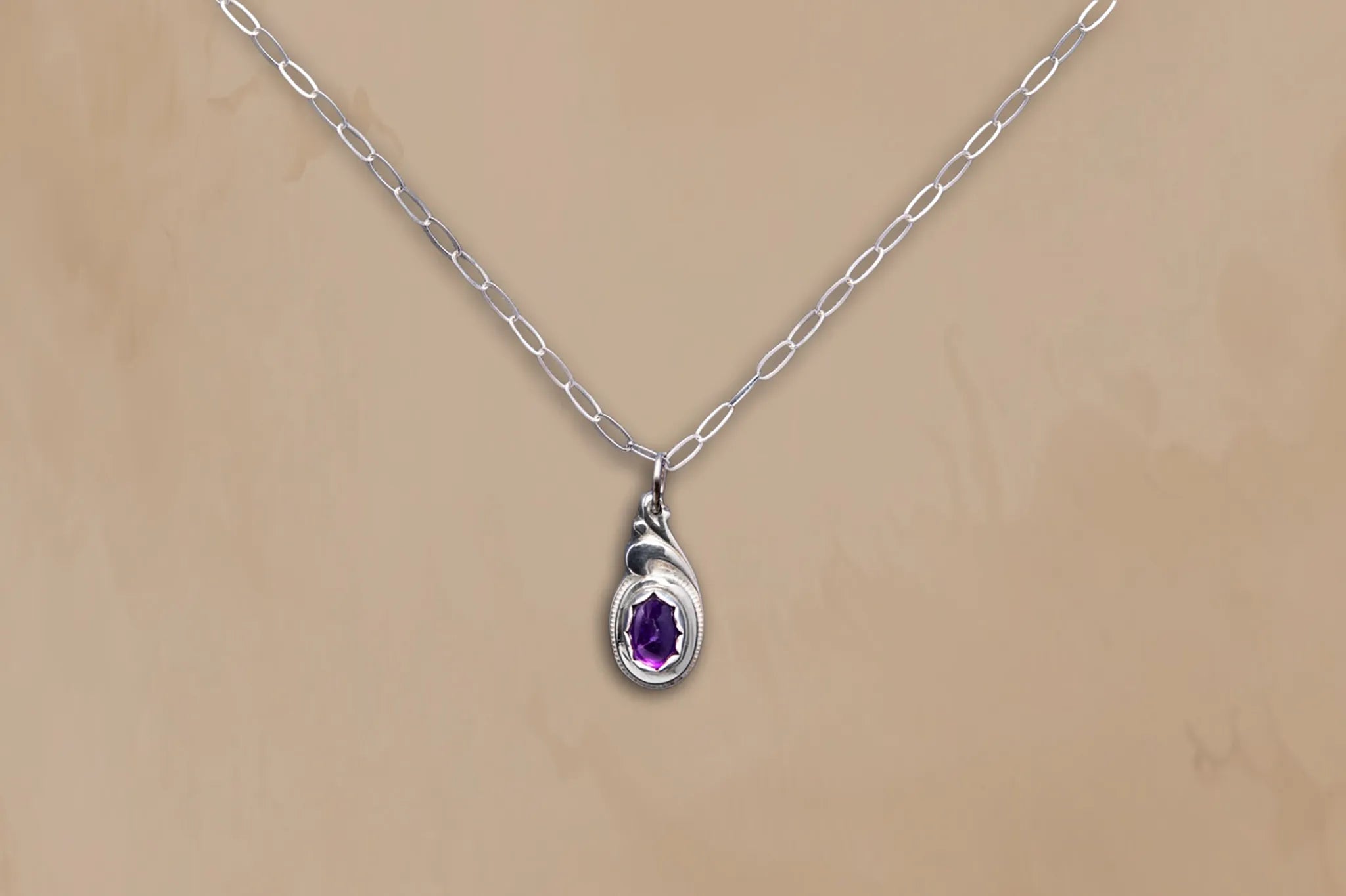 Silver necklace with a teardrop-shaped pendant featuring a purple gemstone on a beige background