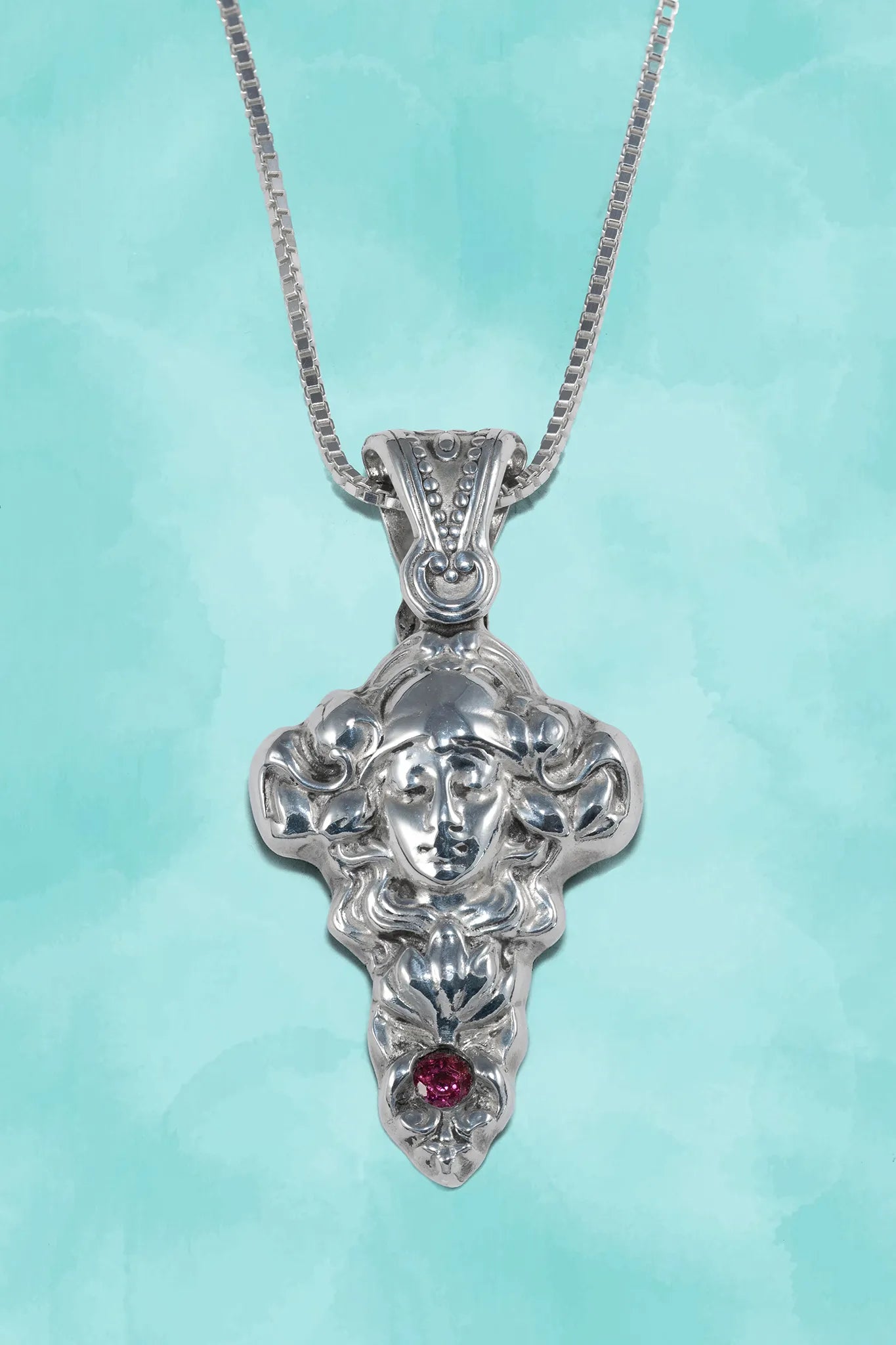 Silver pendant with face design and red gemstone on a light blue background