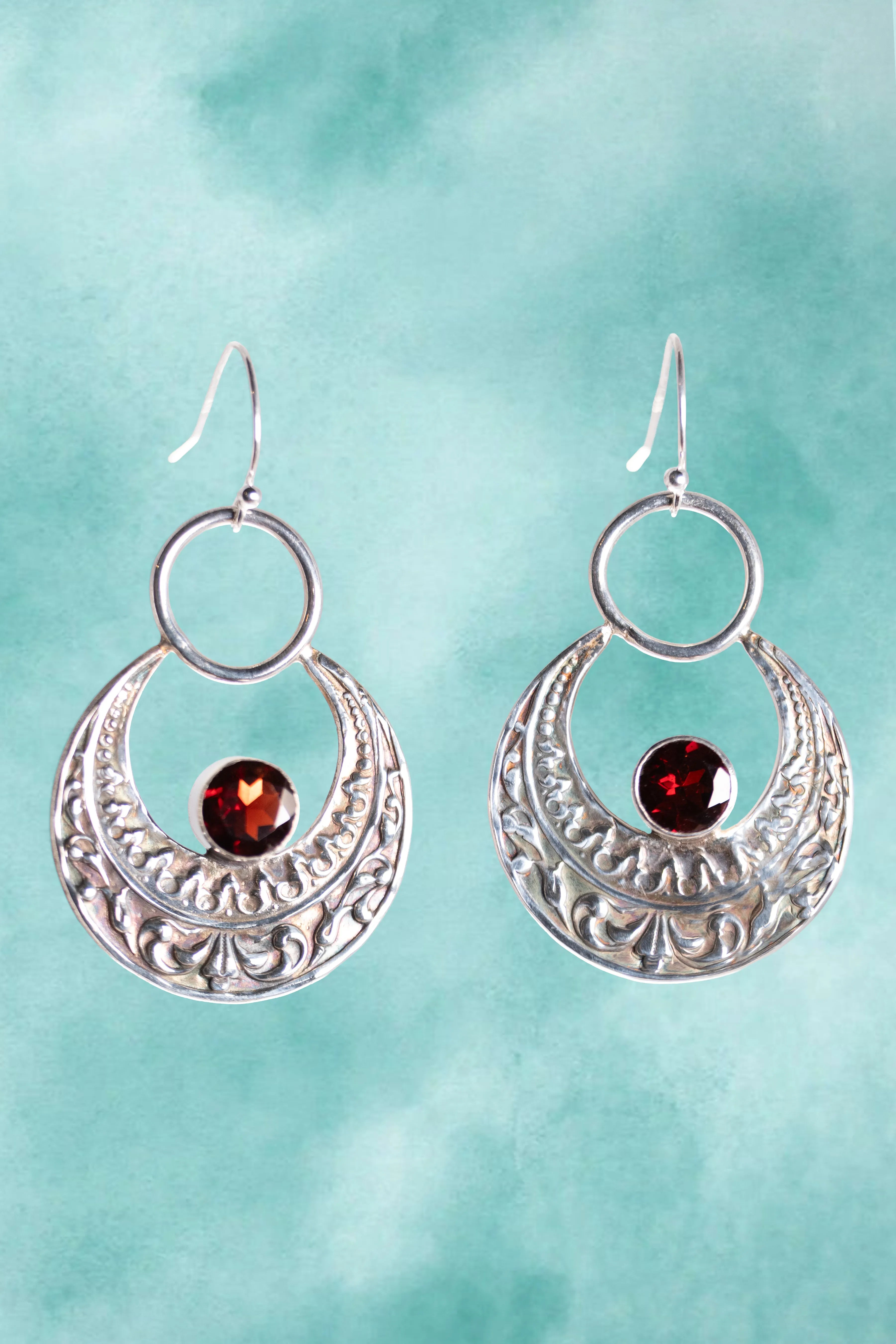 Silver earrings with circular designs and red gemstones on a teal background