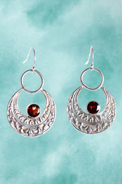Silver earrings with circular designs and red gemstones on a teal background